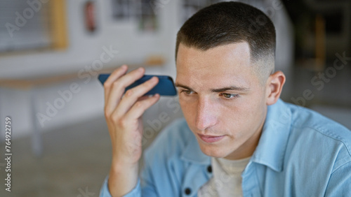 A young hispanic man listens to a voice message indoors, with a focused expression, suggesting a university setting.