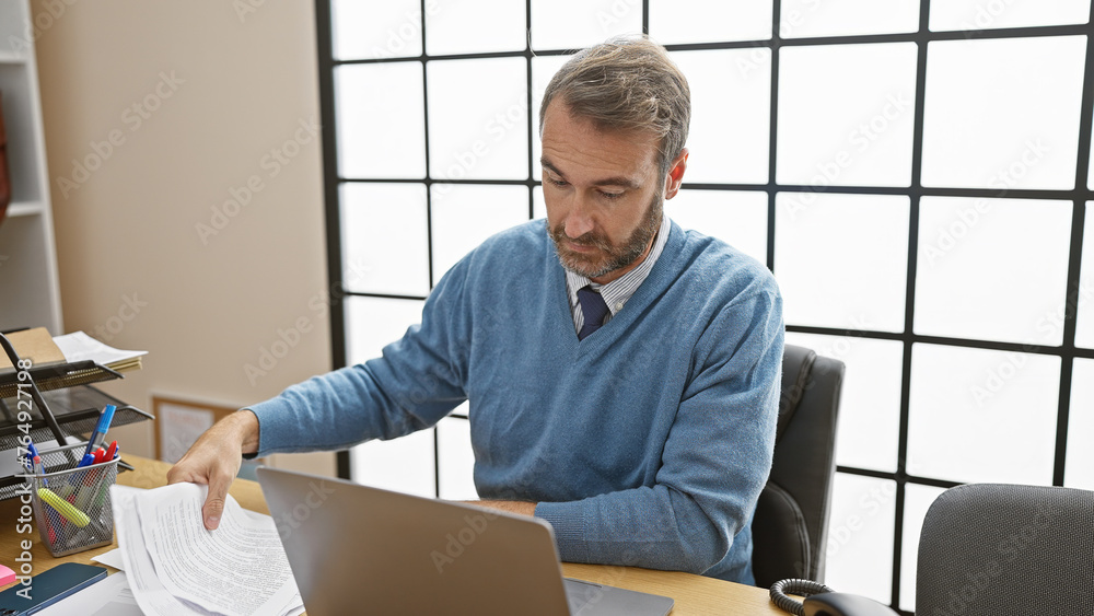 Fototapeta premium Handsome middle-aged hispanic man wearing a sweater and tie working at his office desk on a laptop.