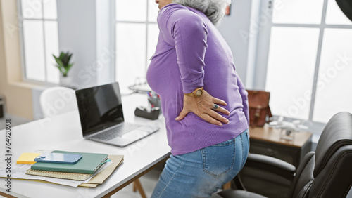 Mature woman in an office setting holding her back, suggesting discomfort or pain.