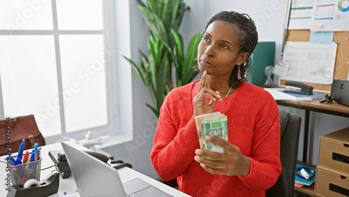 A thoughtful woman in a red sweater holds south african rands in an office, pondering financial decisions. photo