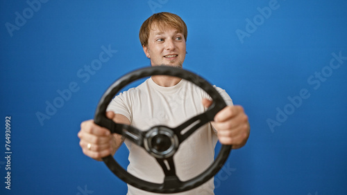 A young, blond man holds a steering wheel against a blue background, suggesting an outdoor or driving concept. photo