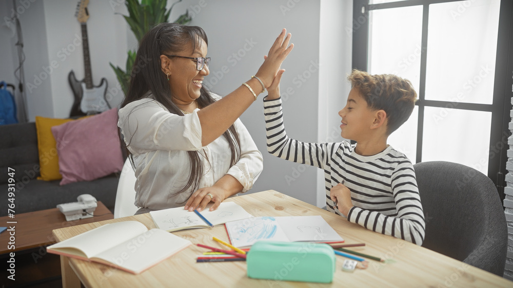 Obraz premium A smiling woman and boy giving high-five at wooden desk with drawings in a well-lit living room, showcasing family bond and happiness.