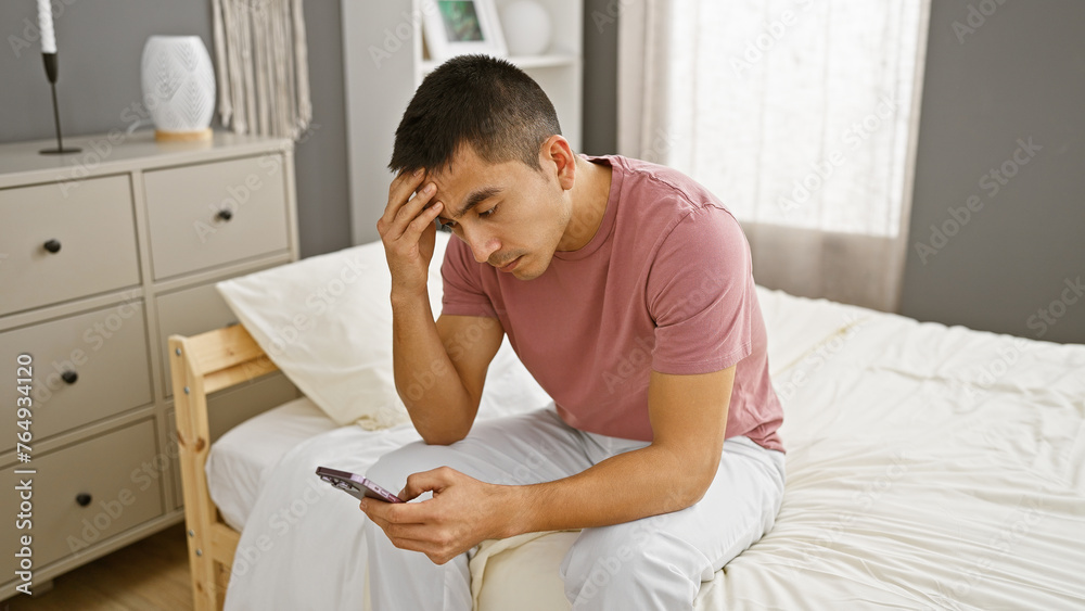 Pensive young hispanic man with phone sitting on bedroom bed in a modern apartment interior. Pensive young hispanic man with phone sitting on bedroom bed in a modern apartment interior.