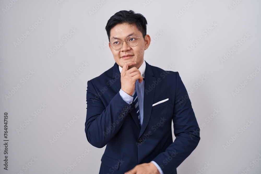 Young asian man wearing business suit and tie with hand on chin thinking about question, pensive expression. smiling and thoughtful face. doubt concept. Young asian man wearing business suit and tie with hand on chin thinking about question, pensive expression. smiling and thoughtful face. doubt concept.