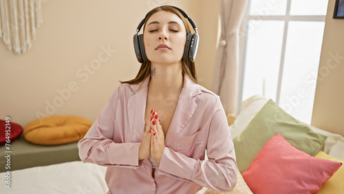 A serene young woman in pink pajamas meditating on a bed with headphones in a cozy bedroom setting. photo