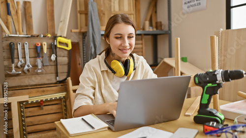 A young woman smiles while using a laptop in a well-equipped woodworking workshop.