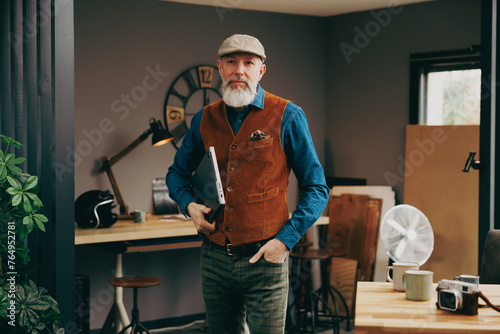 Portrait d'un homme debout souriant quinquagénaire senior hipster élégant et stylé qui fait une pause dans un atelier créatif vintage et qui porte un ordinateur portable photo