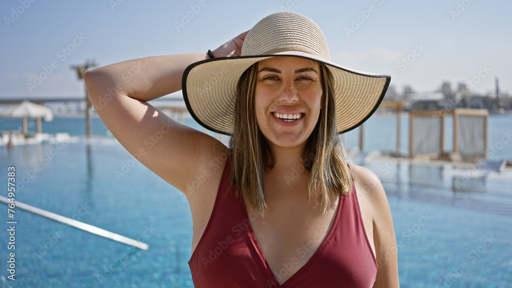 Smiling young hispanic woman with brunette hair, wearing a hat, enjoying luxury poolside at a beach club.