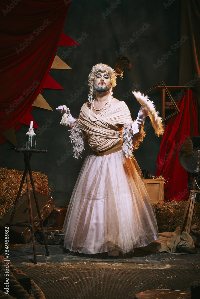Drag queen in white dress and bright makeup over dark retro circus backstage background. Man in female appearance. Concept of circus, theater, performance, show, retro and vintage