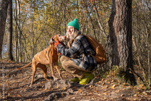 Middle aged happy woman petting vizsla dog walking in nature autumn forest. Female rejoicing spending time with pet friend on weekend sitting on haunches.