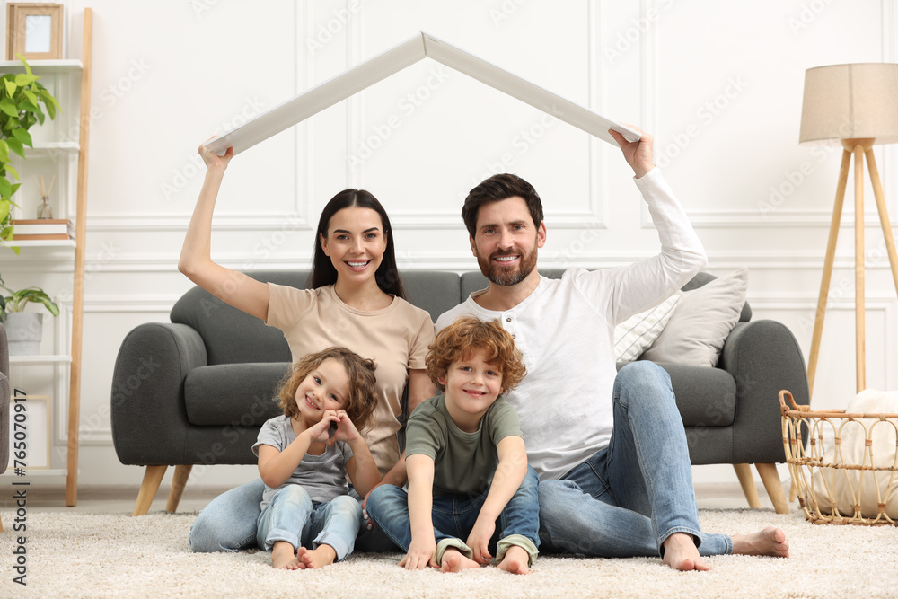 Family housing concept. Happy woman and her husband holding plastic roof while sitting with kids on floor at home Family housing concept. Happy woman and her husband holding plastic roof while sitting with kids on floor at home