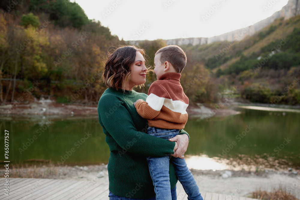 A happy mother holds a four-years-old child in his arms against the background of a lake, forest and mountain A happy mother holds a four-years-old child in his arms against the background of a lake, forest and mountain