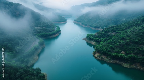 Fog envelops the hills surrounding a tranquil river in a gorge, with verdant green foliage accentuating the peaceful scene.