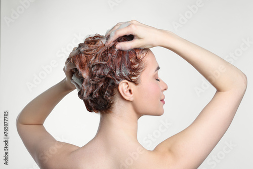 Young woman washing her hair with shampoo on light grey background, back view