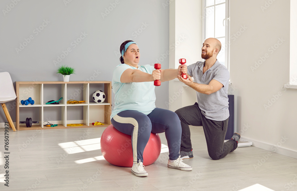 Obraz premium Plus size, fat woman training with dumbbells under guidance of personal trainer in gym. Obese, overweight woman sitting on fitball doing exercise with dumbbells. Weight loss, healthy lifestyle