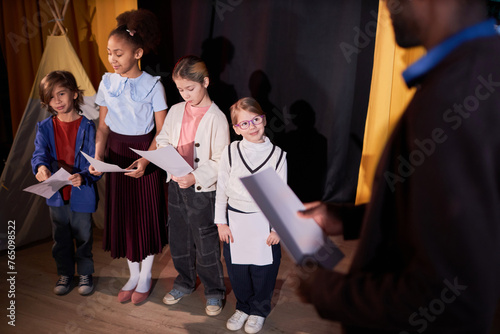 Over shoulder view of school drama teacher directing group of kids rehearsing on stage in theater