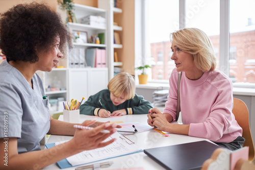Side view portrait of young mother talking to pediatrician in doctors office with child in background
