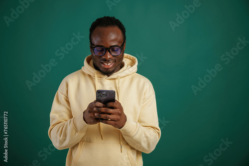 Using smartphone. African American man is standing against green background in the studio