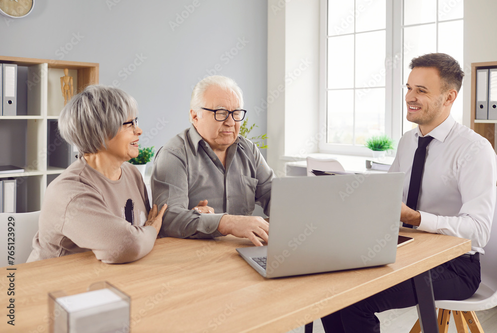 Smiling happy elderly couple talking with man advisor about health insurance or investments sitting at the desk in office with laptop. Senior man and woman talking with male financial agent. Smiling happy elderly couple talking with man advisor about health insurance or investments sitting at the desk in office with laptop. Senior man and woman talking with male financial agent.