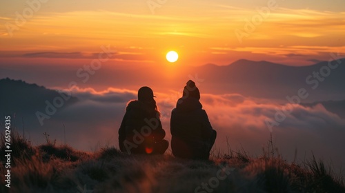 Two friends sitting on a mountaintop watching the sunset. The sky is a bright orange and the sun is just setting below the horizon.