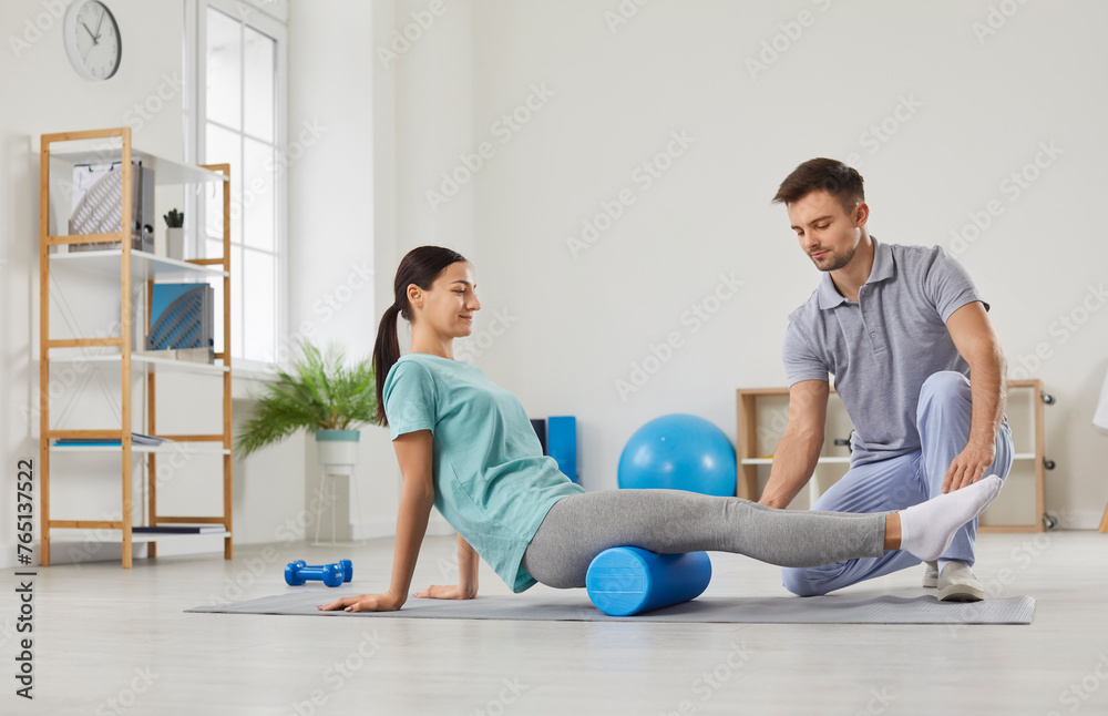Young smiling woman patient doing exercise using sport massage roller with support from male physiotherapist doctor in rehab clinic. Physiotherapy and physical rehabilitation concept. Young smiling woman patient doing exercise using sport massage roller with support from male physiotherapist doctor in rehab clinic. Physiotherapy and physical rehabilitation concept.