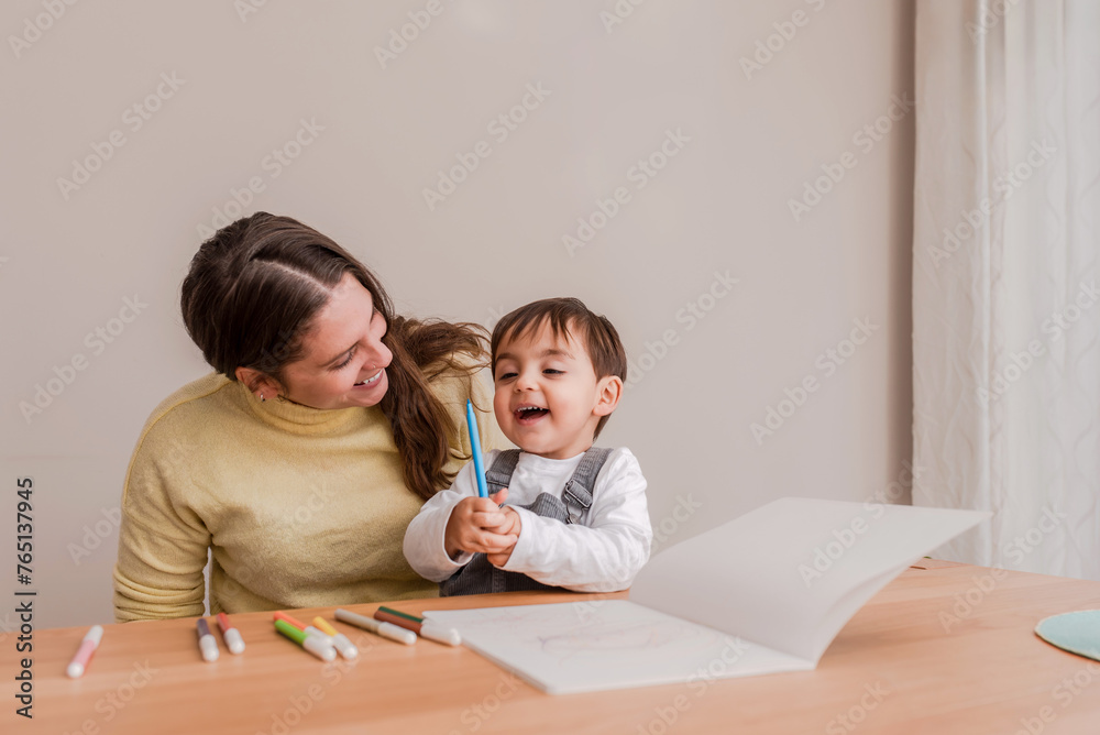 Happy Young toddler Boy Drawing with Markers sitting on the table with his smiling mother at Home Happy Young toddler Boy Drawing with Markers sitting on the table with his smiling mother at Home