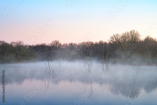 A small lake at sunrise covered with fog from the cold night. There are old dead trees in the lake. There are nice reflections in the water. 