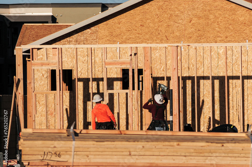 In a housing development in Queen Creek, Arizona, two wood framers wearing hard hats are using power equipment, such as a pneumatic nailer, to construct the wooden framework of a tract home