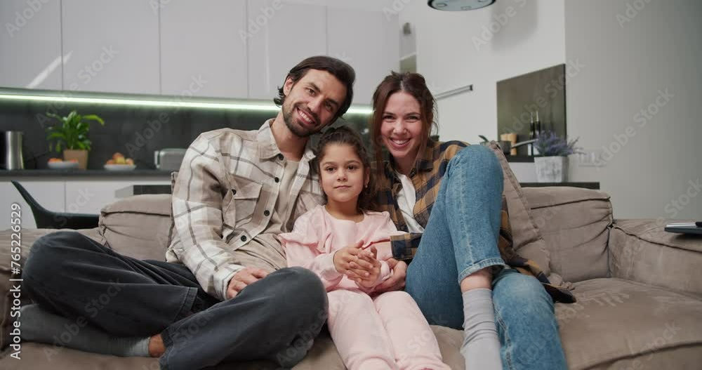 Portrait of a happy family, a brunette man with stubble along with his brunette wife in a checkered shirt sitting on a brown sofa along with their little daughter in a pink suit in a modern new Portrait of a happy family, a brunette man with stubble along with his brunette wife in a checkered shirt sitting on a brown sofa along with their little daughter in a pink suit in a modern new