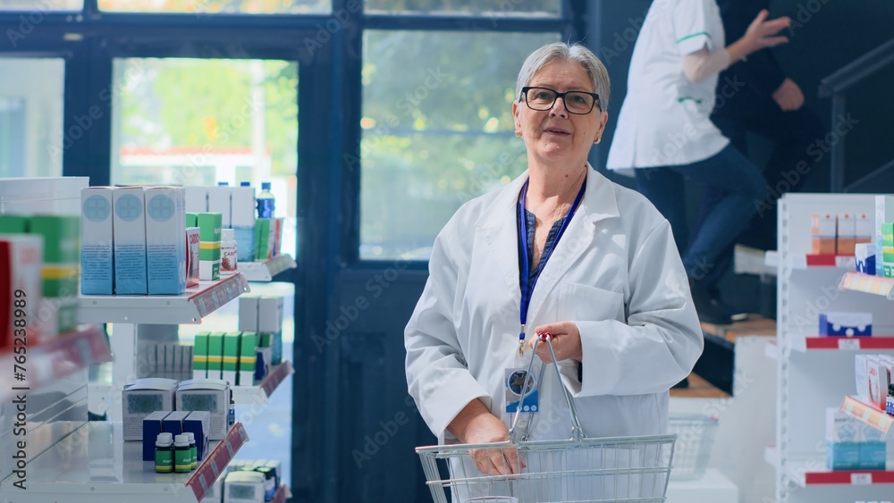 Aged knowledgeable druggist in pharmacy holding shopping basket, arranging merchandise on shelves. Octogenarian pharmacist organizing medicinal products in drugstore with clients in background Aged knowledgeable druggist in pharmacy holding shopping basket, arranging merchandise on shelves. Octogenarian pharmacist organizing medicinal products in drugstore with clients in background