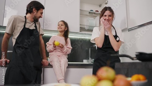 Happy brunette girl talking on the phone in a white T-shirt and black apron together with her brunette husband with stubble communicate and have fun with their little daughter in pink clothes while
