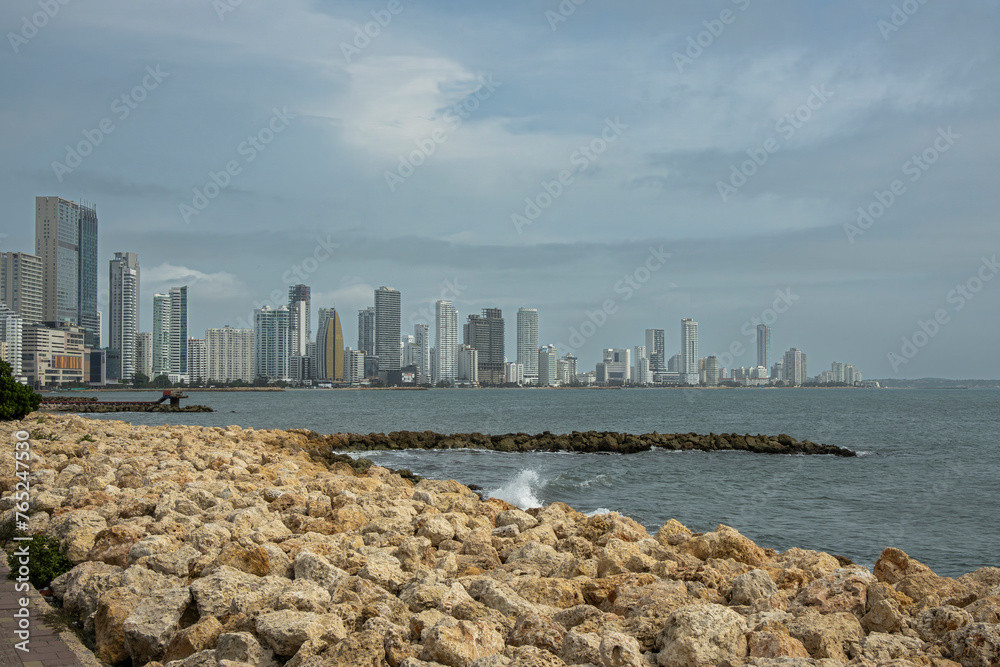 Naklejka premium Cartagena, Colombia - July 25, 2023: Seen from where Calle 33 meets Avenida Santander, breakwater and Tall buildings skyline on NW side of Bocagrande peninsula. Rocky shore upfront