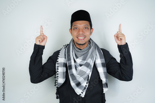 Cheerful handsome Asian Muslim man wearing Arab turban sorban pointing hand finger at empty space for advertising on isolated background photo