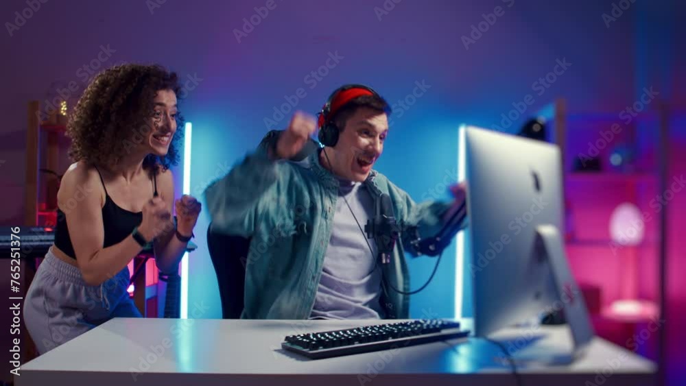 Excited Gamer Guy and Young Woman wearing headphones playing computer game neon fashion room, winner. Gamer winning hard match, guy's girlfriend is happy and congratulates her boyfriend looking at Excited Gamer Guy and Young Woman wearing headphones playing computer game neon fashion room, winner. Gamer winning hard match, guy's girlfriend is happy and congratulates her boyfriend looking at