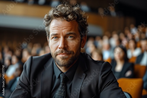 A man in a suit and tie, with a beard and facial hair, is sitting in front of a crowd at a formal event. He looks pleased, adjusting his collar and gesturing with confidence