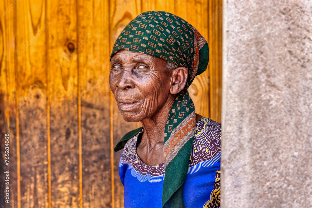 village african old woman portrait, she is standing in front of the door and wearing a green scarf and blue dress