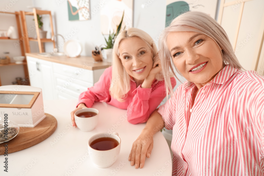 Mature female friends with tea taking selfie at table in kitchen