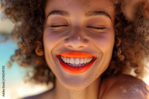 Beautiful young woman with curly brown hair and bright orange lipstick smiling under the warm sun with closed eyes, expressing joy and happiness