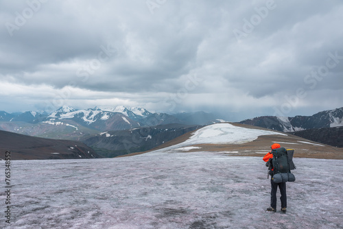Man on big glacier with panoramic view from snow rock hill to large snow-capped mountain range in rainy low clouds. Guy in high snowy mountains under gray cloudy sky. Dramatic alpine vast landscape.