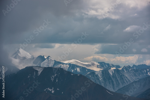Dark atmospheric landscape with mountain silhouettes and large snow-capped peaked top in rainy low clouds. Dramatic alpine view to high mountains in gray cloudy sky. Big snowy mountain range in rain. © Daniil