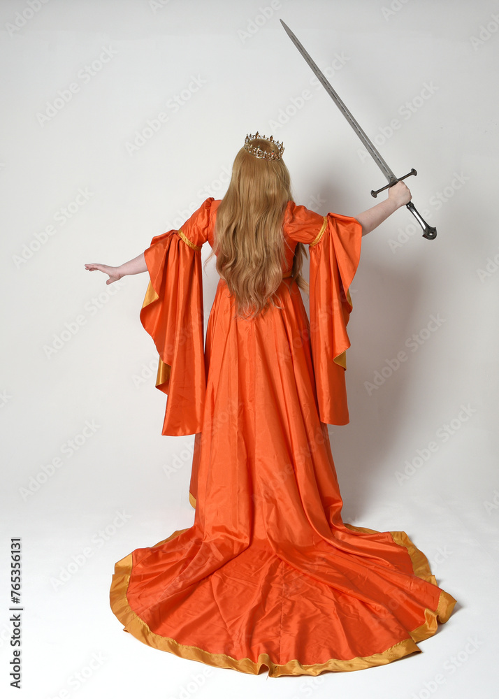 Obraz premium Full length portrait of plus size blonde woman, wearing historical medieval fantasy gown, golden crown royal queen. Standing pose backview, holding sword weapon, isolated studio background.