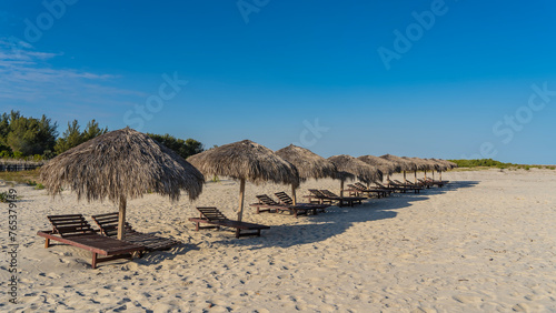 Wooden deck chairs stand in a row under sun umbrellas. Shadows on the sand. Footprints. Green vegetation in the distance. Clear blue sky. Copy space. Madagascar. Morondava.