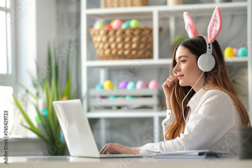 Side view portrait of businesswoman sitting at the home office desk and looking at laptop screen. She is wearing bunny ears headphones. Colorful easter eggs in the background. © zphoto83