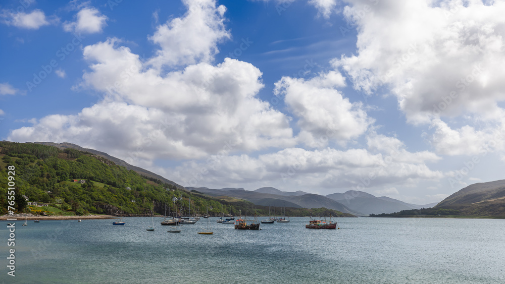 Fishing boats anchors in the gentle waters of Ullapool, set against the stunning vista of the Scottish Highlands under a sky dotted with clouds