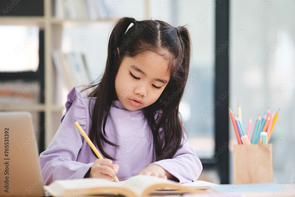 A young girl is sitting at a desk with a book and a laptop A young girl is sitting at a desk with a book and a laptop