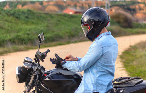 Young man putting on gloves on top of motorbike with le helmet on and road with landscape in background, casual dress