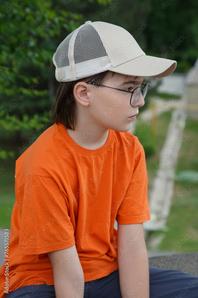 A boy in a cap and orange against a background of greenery outside.