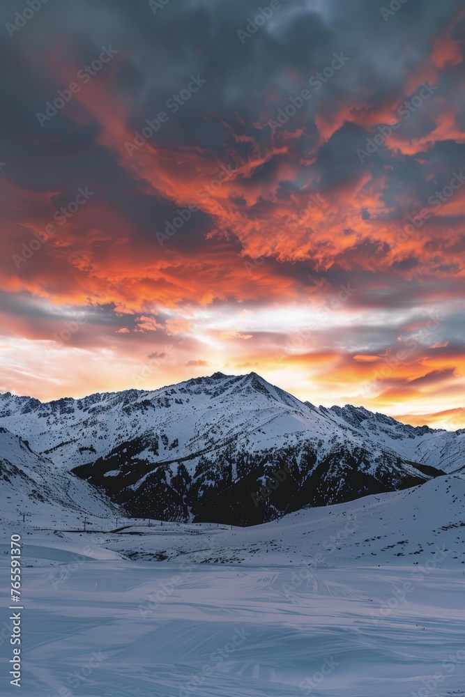 The sun sets over a snowy mountain range, casting warm hues across the clouds in the sky