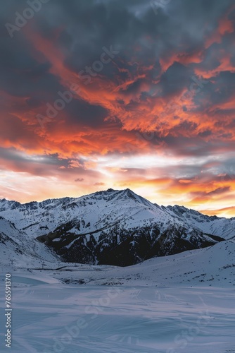 The sun sets over a snowy mountain range, casting warm hues across the clouds in the sky