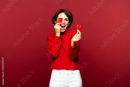 Cheerful, happy woman smiling, holding small hearts, covering one eye, isolated on a red background 